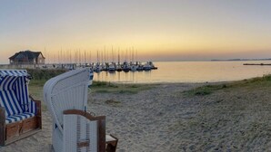 Une plage bordée d’une rangée de chaises en osier blanc et d’une chaise à rayures bleues, une structure en bois et plusieurs bateaux amarrés à un quai.