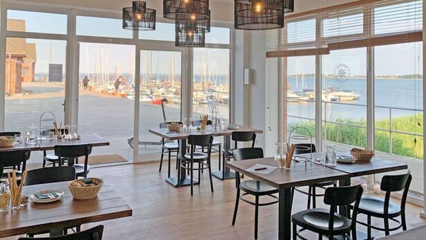 Un espace repas doté de grandes fenêtres donnant sur un port de plaisance, avec des tables et des chaises en bois, ainsi que des lampes suspendues.