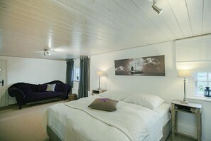 Een slaapkamer met een bed, een nachtkastje, een lamp, een raam, een schilderij aan de muur en een paarse bank.