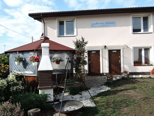 Una casa de dos pisos con un cartel que dice "Szaffirowe Zaciszce" y un número de contacto. Hay un pequeño jardín con un bebedero para pájaros y plantas en macetas.