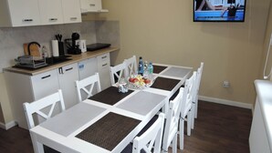 A dining area with a table set for four, surrounded by white chairs. A television is mounted on the wall, displaying a program. - Holiday House in Wapnica Near Emerald Lake (Międzyzdroje)