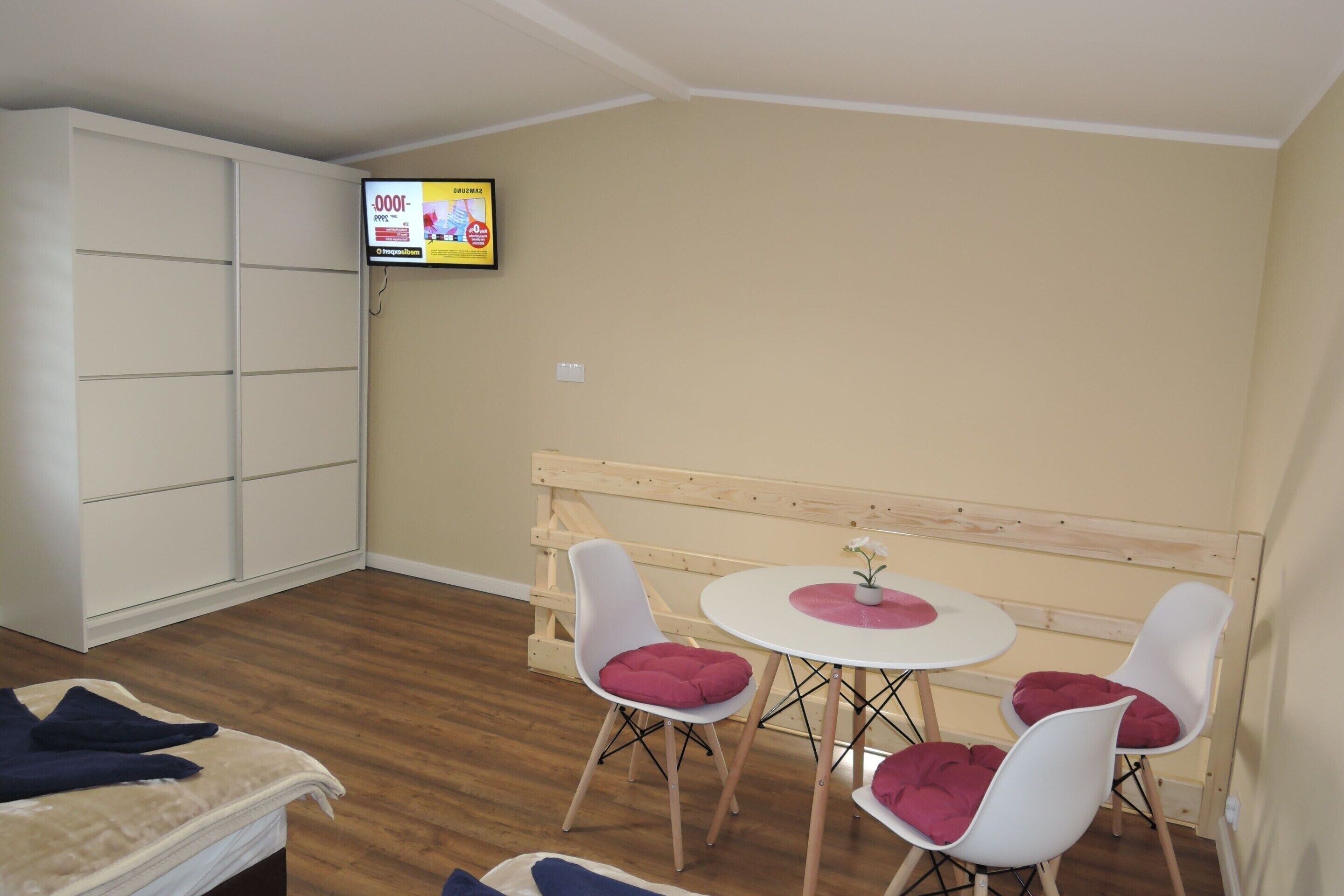 Ein kleines Zimmer mit einem Bett, einem an der Wand befestigten Fernseher, einer hölzernen Bank und zwei weißen Stühlen mit roten Kissen.