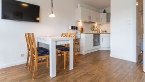 A modern kitchen with white cabinets, a dining table with chairs, and a flat-screen TV mounted on the wall. - Ferienwohnung, St. Peter-ording (St. Peter-Ording)