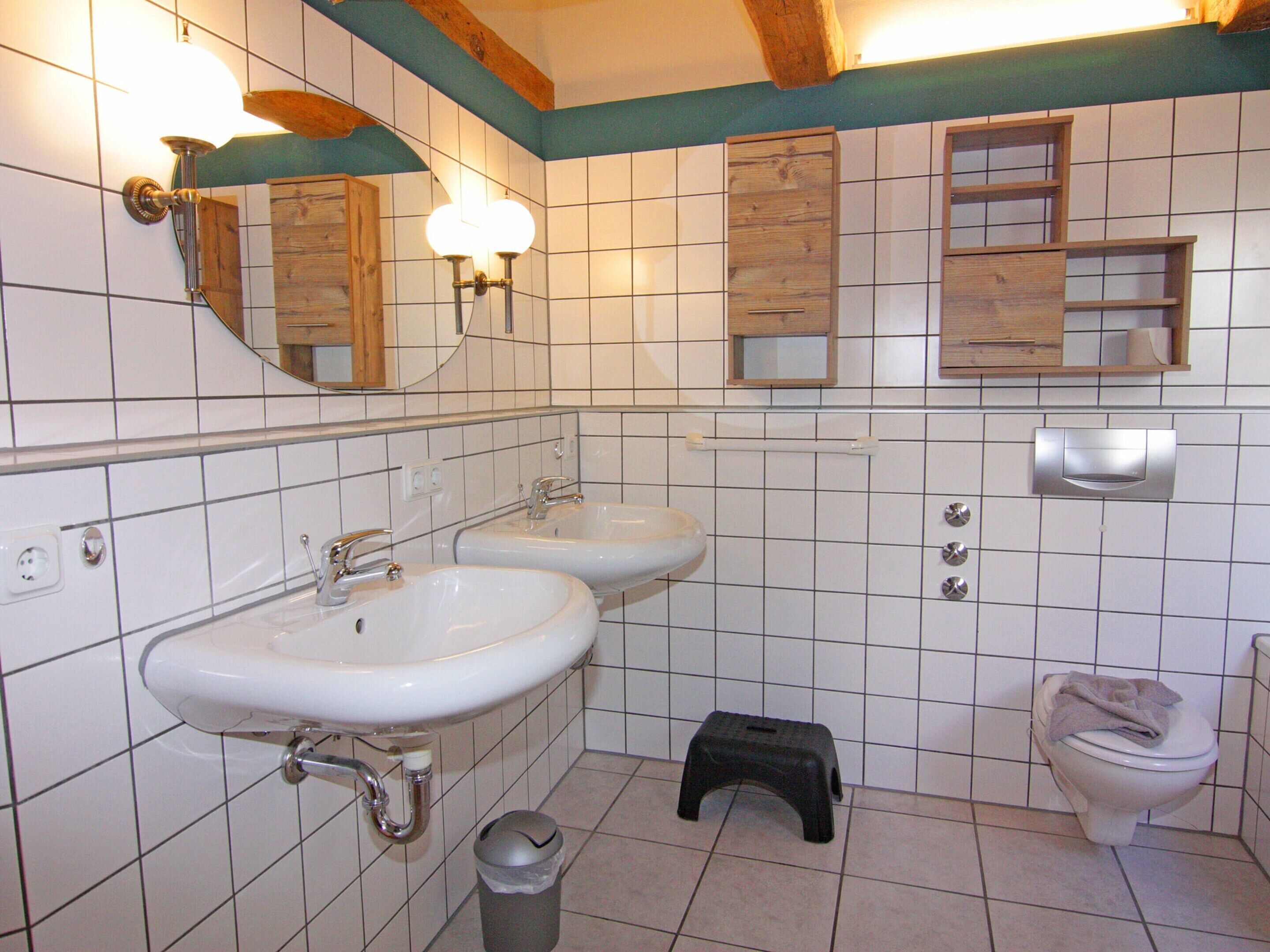 A bathroom with two sinks, a toilet, a trash bin, and wooden cabinets above the sinks.