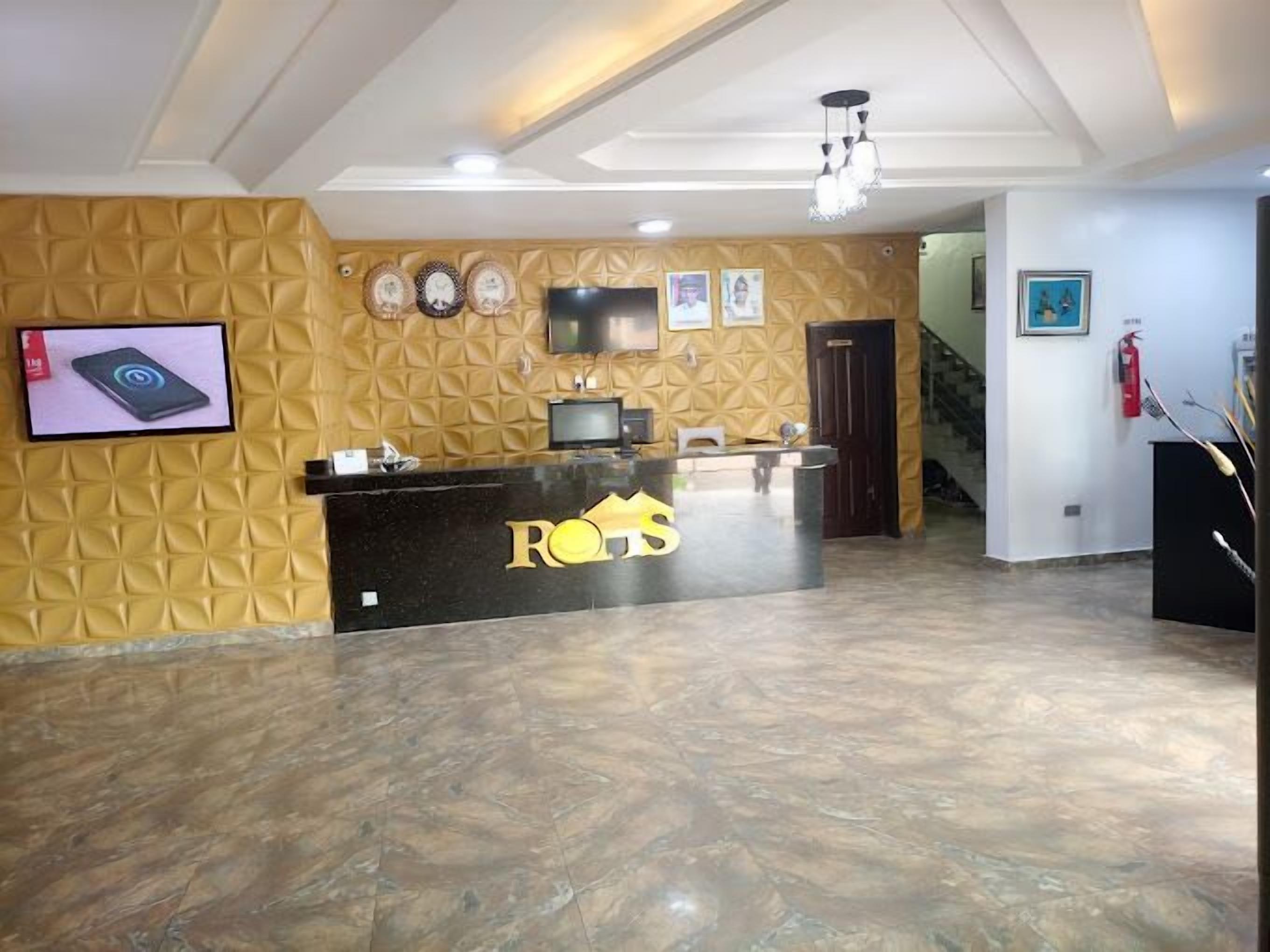 Un área de recepción con un mostrador negro que muestra el logo 'ROS'. Hay dos televisores de pantalla plana montados en la pared, un accesorio de iluminación en el techo y un extintor a la derecha.