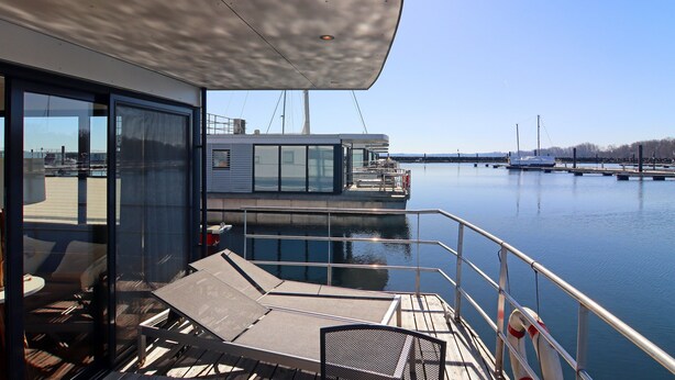 A modern deck with lounge chairs and a table, overlooking calm water and a marina with boats.