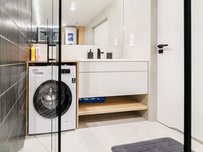 Et lille vaskerum med en Samsung vaskemaskine, en hylde med toiletartikler og et spejl over en vask.