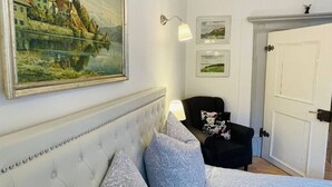 Una camera da letto con testiera trapuntata, un letto con biancheria a motivi, una poltrona nera con cuscino floreale, una lampada a parete e quadri appesi.