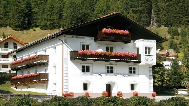 A multi-story building with balconies adorned with flowers, a sign indicating it is an apartment hotel, and a mountainous backdrop.