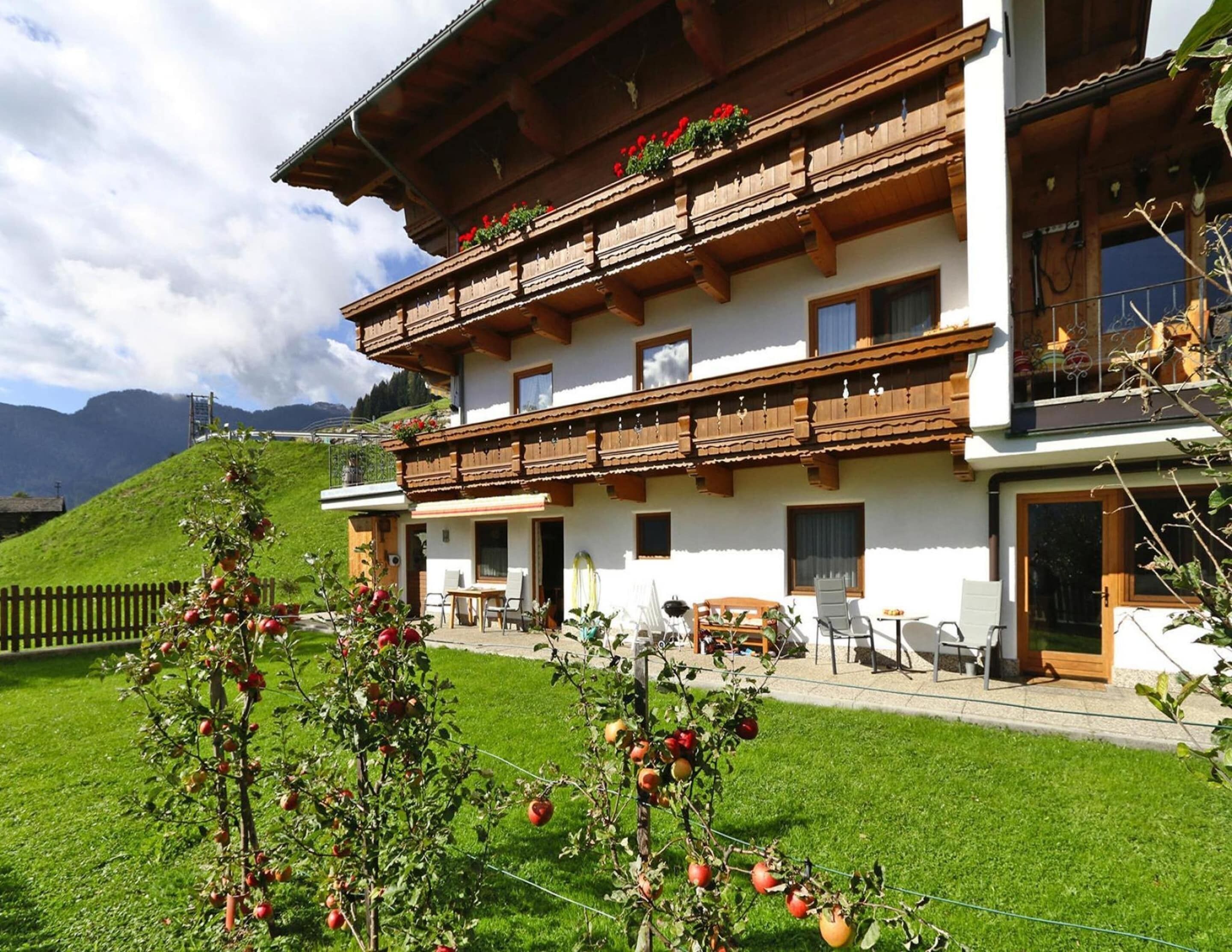 A two-story chalet with wooden balconies, a garden with apple trees, and a mountain backdrop.