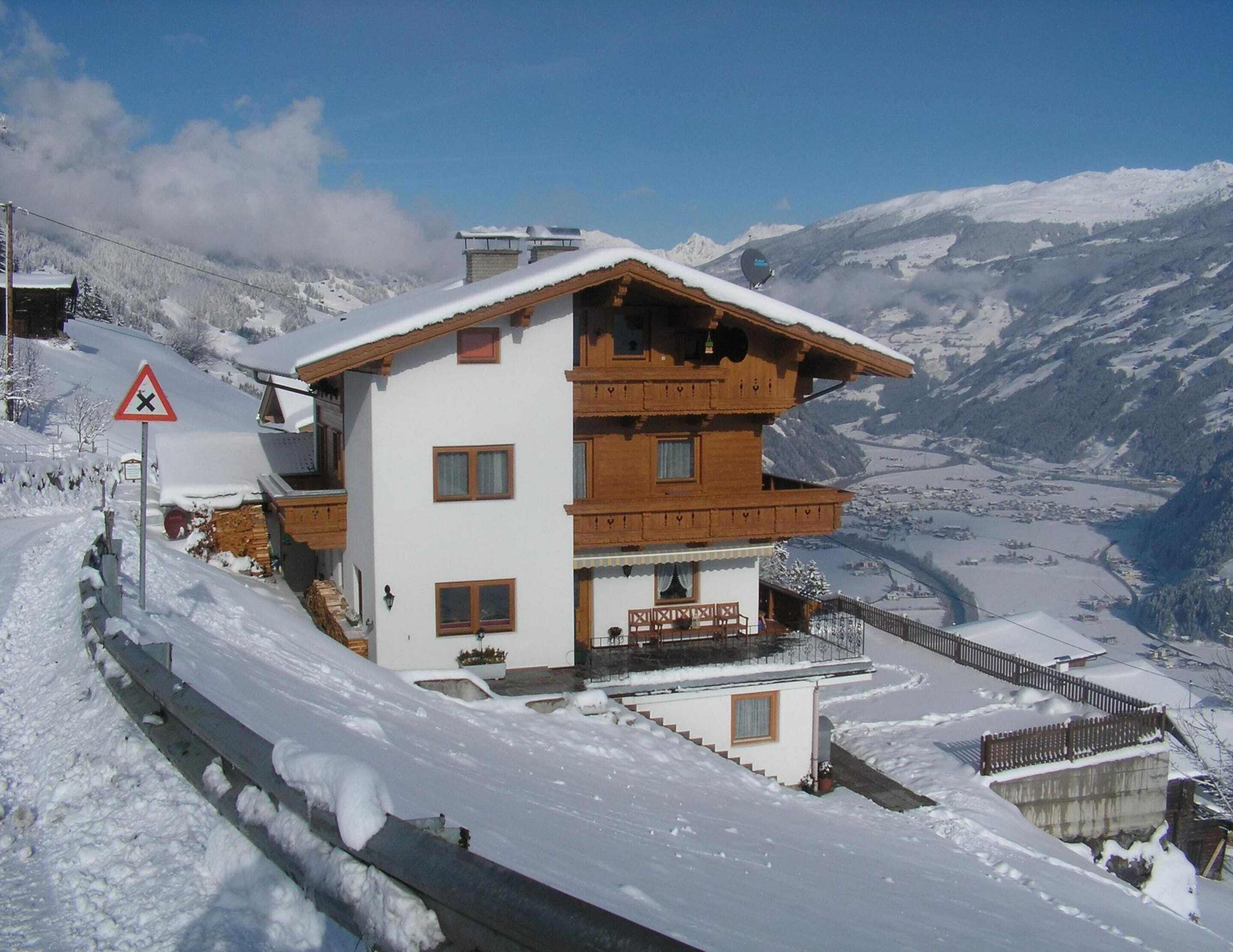 A two-story wooden chalet with a balcony, surrounded by snow-covered mountains.