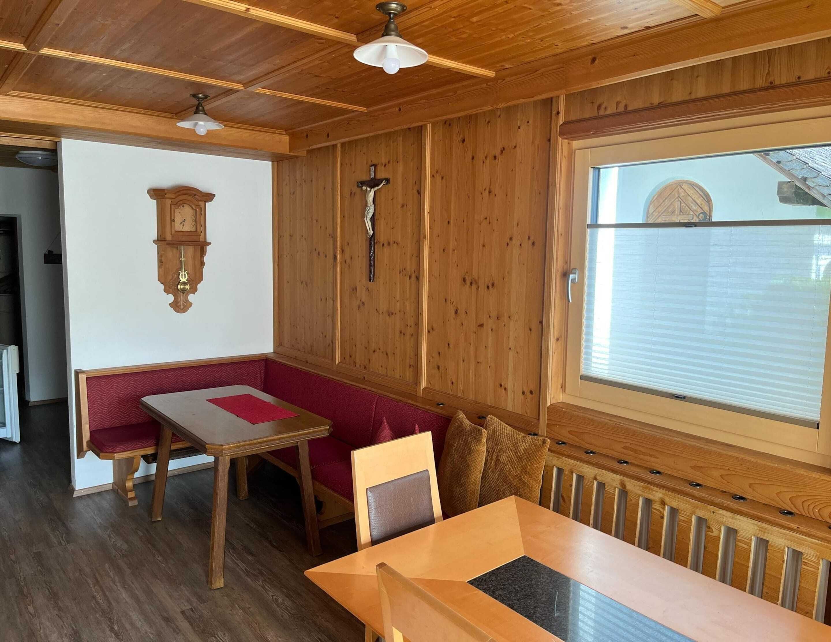 A cozy cabin interior with a wooden ceiling, a dining table with red cushions, a wall-mounted clock, and a window with blinds.