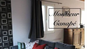 A modern living room with a grey sofa, a matching ottoman, and a patterned wall. - Un Séjour Nature au Coeur de L'histoire (Nonant)