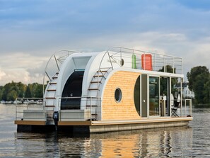 A houseboat with a wooden deck, a glass-enclosed lounge area, and a small outdoor seating area. - Houseboat on the Dahme (Königs Wusterhausen)