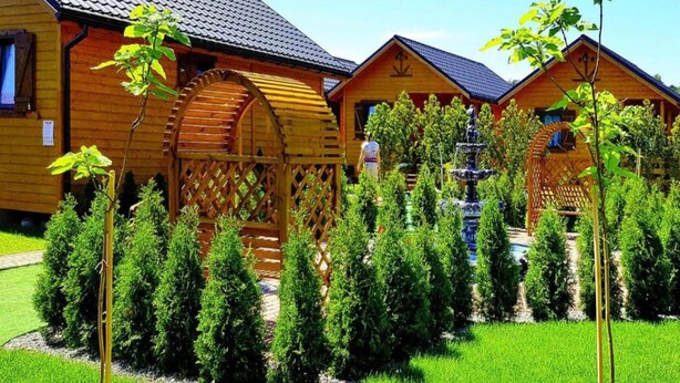A wooden cabin with a large water wheel, surrounded by a well-maintained garden with trimmed bushes and a lawn. - Chalet by Baltic Sea Near Wiselka Lake (Międzyzdroje)