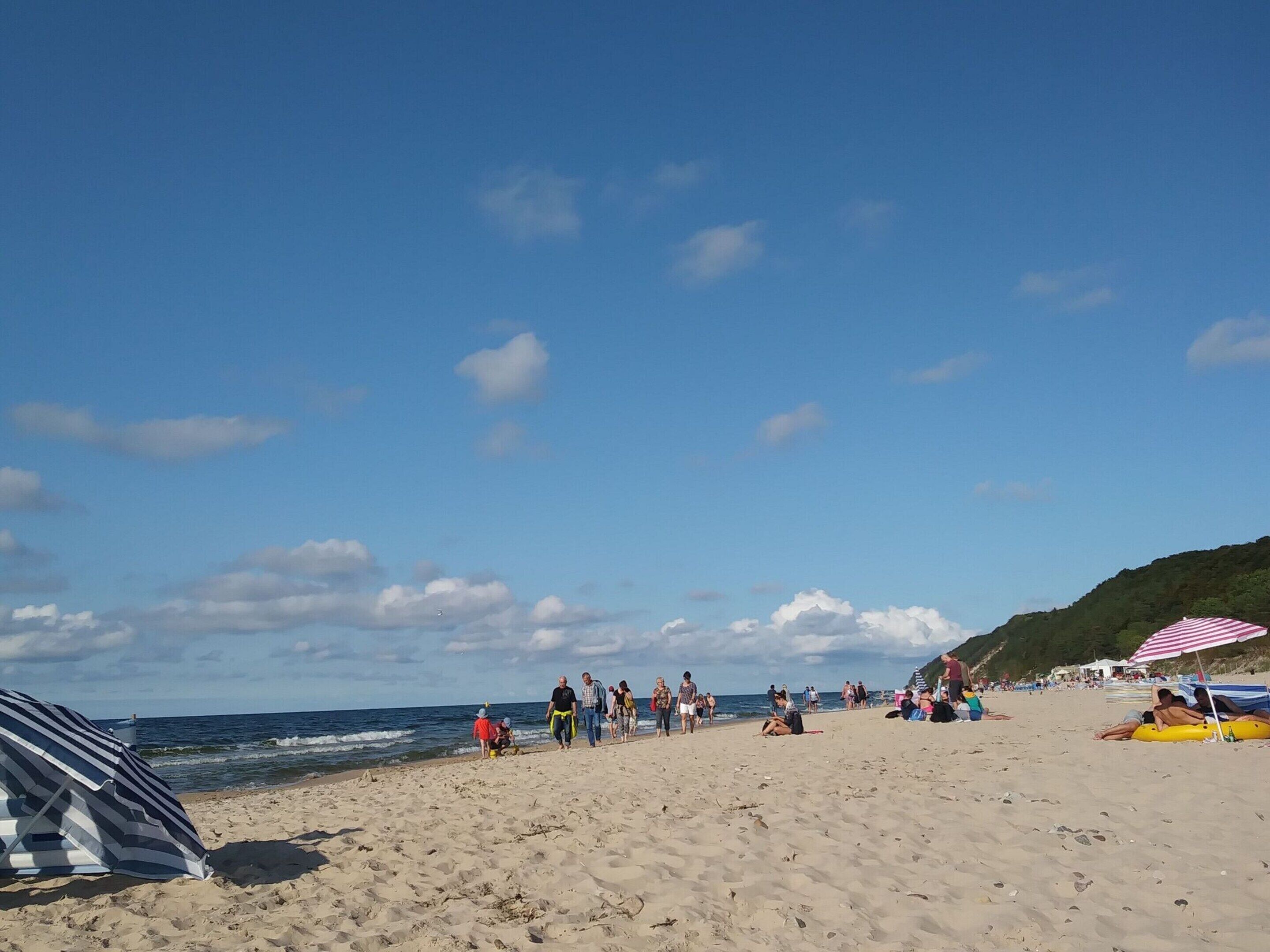 Uma praia arenosa com pessoas caminhando e sentadas, um guarda-sol listrado e uma colina ao fundo.