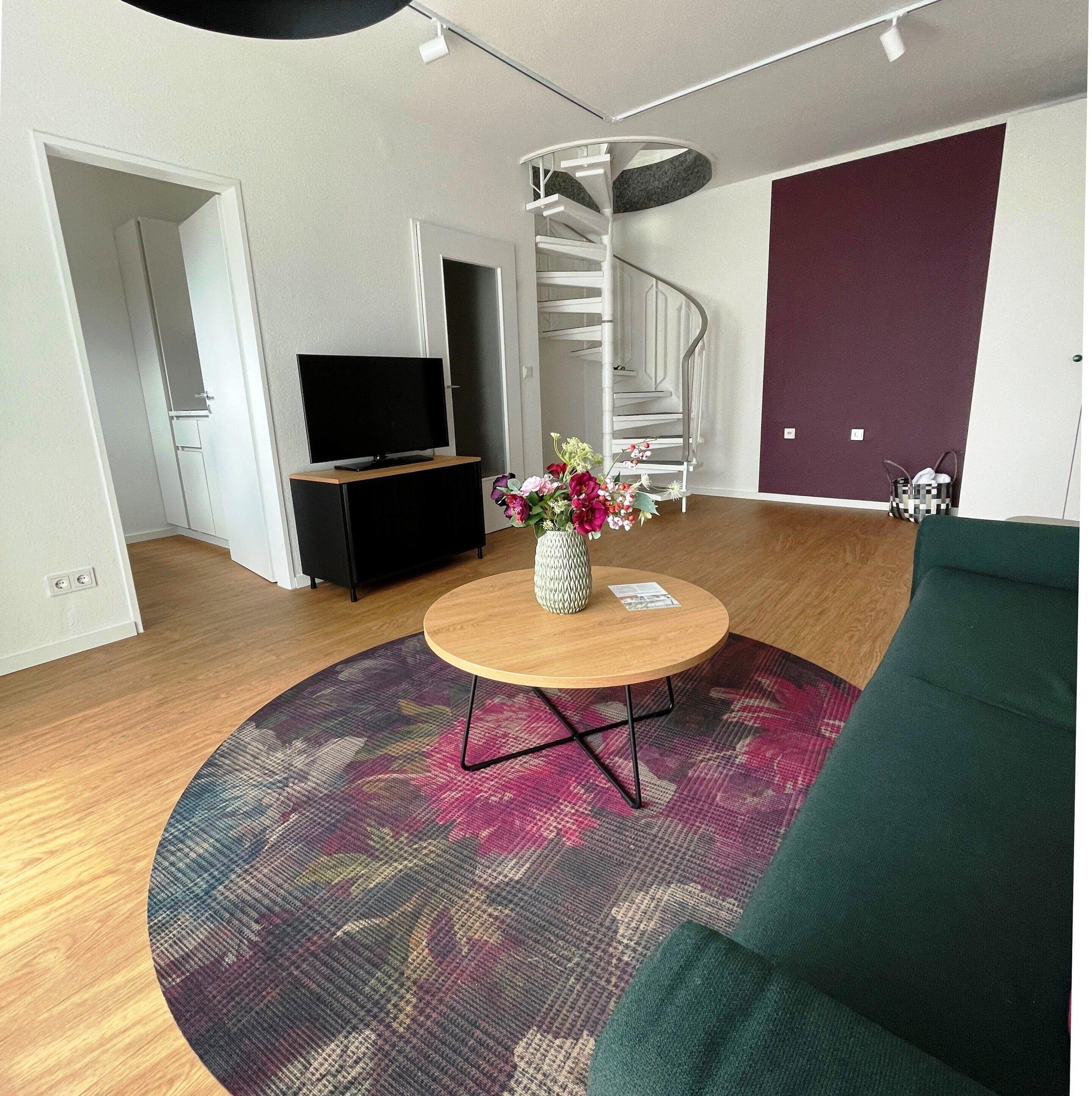 Een moderne woonkamer met een flatscreen televisie, een ronde houten salontafel met een bloevas, een spiltrap en een groene bank.