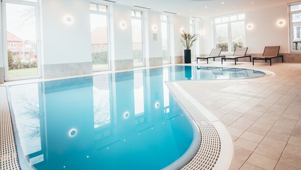 A modern indoor swimming pool with clear blue water, surrounded by large windows and a few lounge chairs. - Komfortable Wohnung mit SPA und Pool (Langeoog)