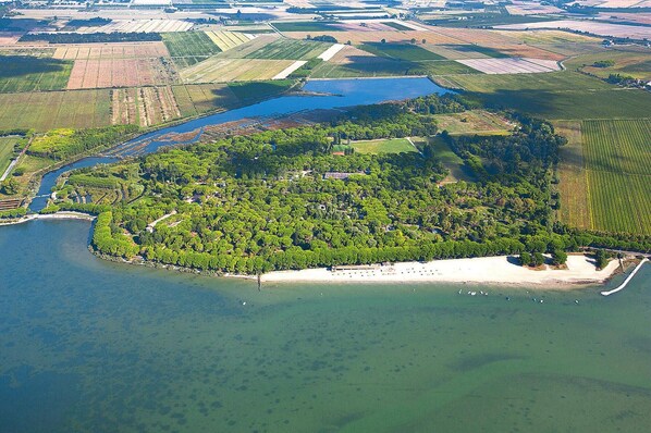 An aerial view of a coastal area with a sandy beach, a small island with dense greenery, and fields in the background. - Grado Belvedere AC Apartment (Aquileia)
