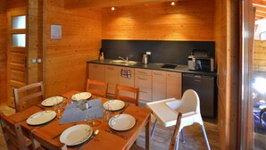 Interno di una baita in legno con un tavolo da pranzo per quattro persone, un angolo cottura dotato di elettrodomestici moderni e una porta vetrata che dà all'esterno.