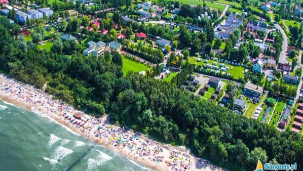 Una vista aerea di una cittadina costiera con una spiaggia, zone residenziali e campi verdi.