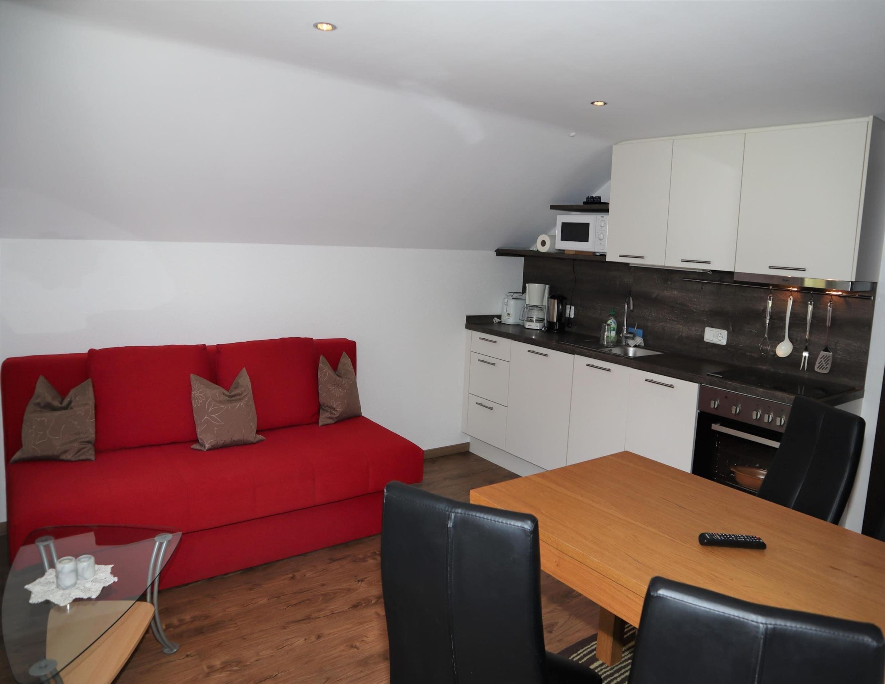 A compact living space with a red sofa, a wooden dining table, and a kitchenette with white cabinets and a microwave.