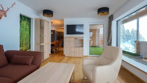 A modern living room with a sofa, a coffee table, a TV, and a large window with a view of trees. - Kitzbuhel Alpine Lodge With Private Infrared Sauna (Mittersill)