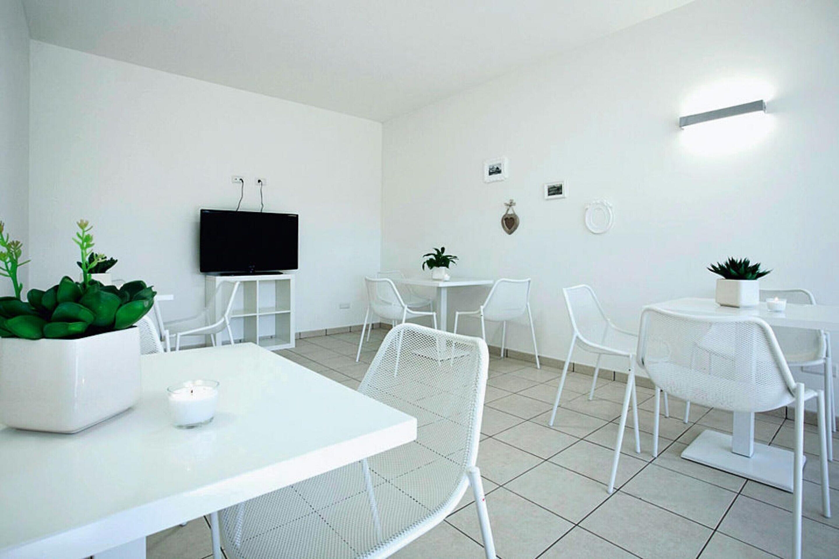 Una camera moderna con arredamento bianco, dotata di TV a schermo piatto montata a parete, sedie e tavoli disposti per mangiare o socializzare.