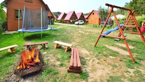 Une cabane en bois avec un trampoline, une balançoire et un foyer au milieu d’une cour gazonnée.