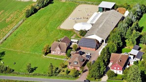 Vue aérienne d’une propriété rurale comprenant plusieurs bâtiments, notamment une grande grange, des maisons plus petites et un espace clôturé où sont garées des voitures.