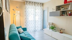 Una sala moderna con un sofá verde azulado, una consola blanca con objetos decorativos y un ventanal con cortinas translúcidas.