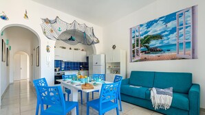A modern kitchen with white cabinets, a dining area with blue chairs, and a teal sofa with a beach view painting. - Casa Celeste 100m From Sea (Porto Cesareo)