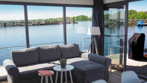 Un espacio habitable moderno con sofá, dos sillones, una mesita y una lámpara. Amplias ventanas ofrecen vistas a un cuerpo de agua y un litoral rocoso.