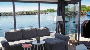 Un área de estar moderna con un sofá, una pequeña mesa y una silla, con vista a un cuerpo de agua con un muelle y un bote.