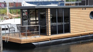 A modern boat with a wooden deck, glass windows, and a covered seating area. - Hausboot Leni, Flensburg (Flensburg)