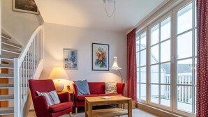 A living room with a red sofa, a wooden coffee table, and a red armchair. There is a staircase on the left and a large window on the right. - Ferienwohnung in St. Peter-ording (St. Peter-Ording)