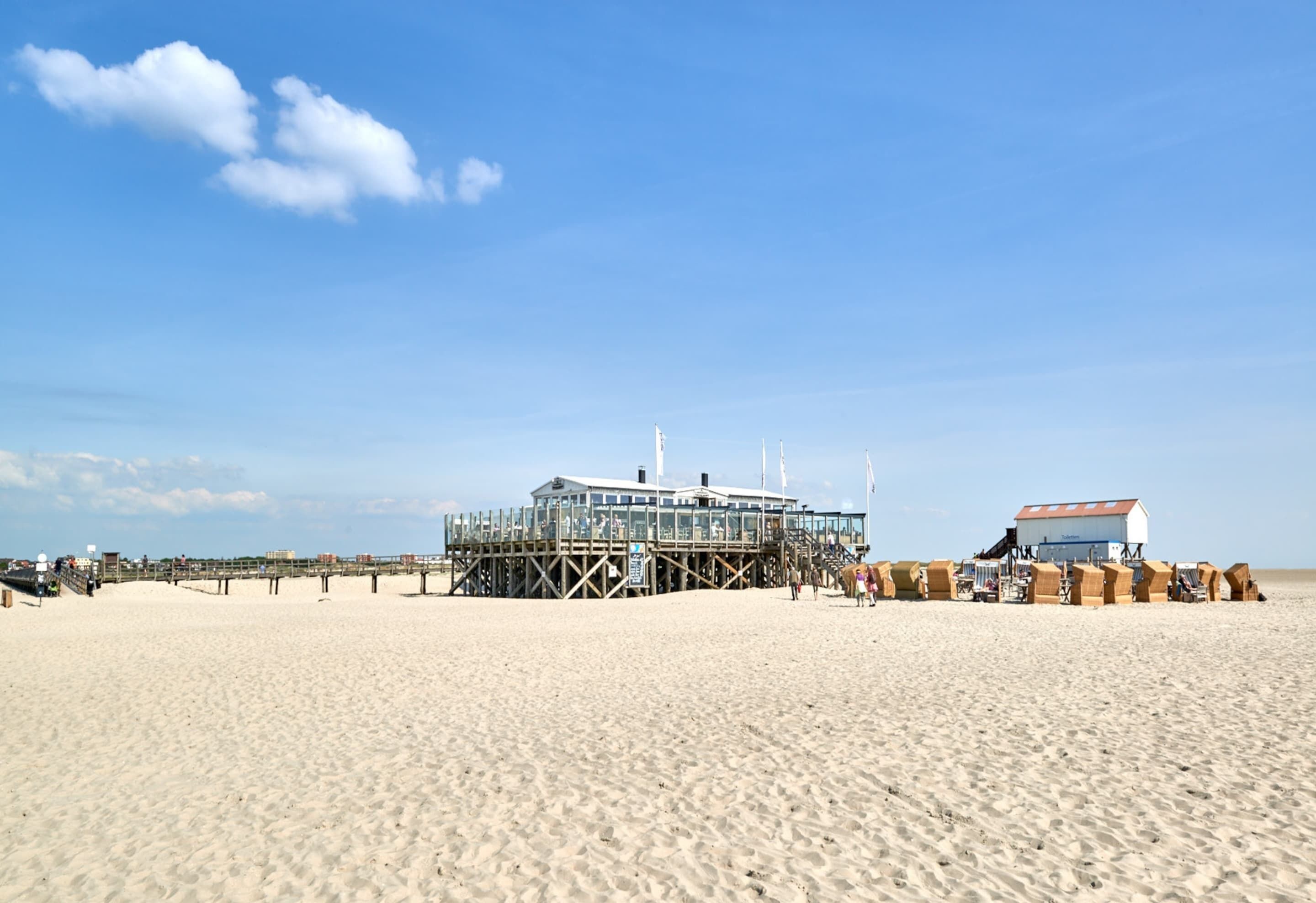 A beach with a wooden pier leading to a glass-enclosed building, likely a restaurant or bar, and a smaller building with a red roof.