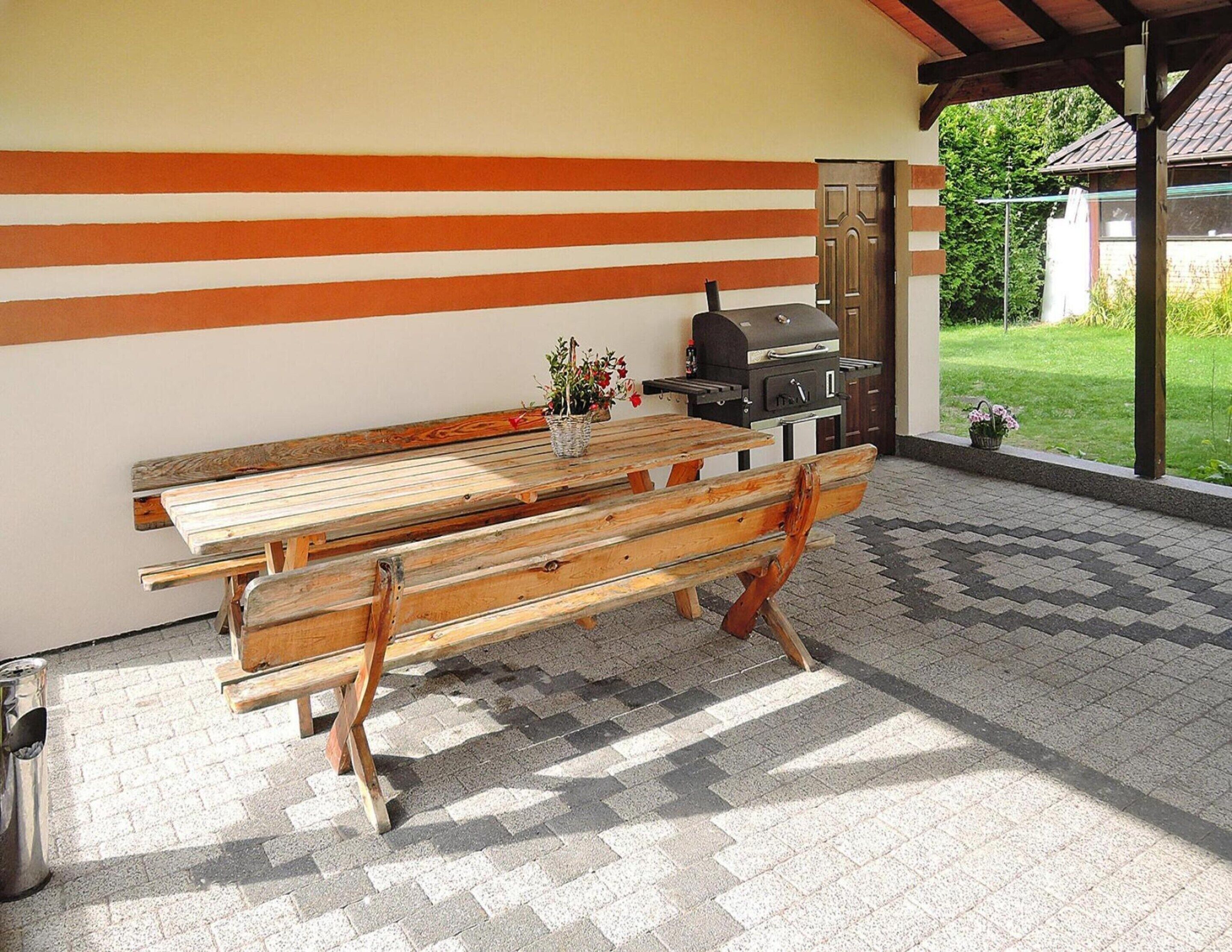 An outdoor patio with a wooden table and benches, a barbecue grill, and a view of a garden.