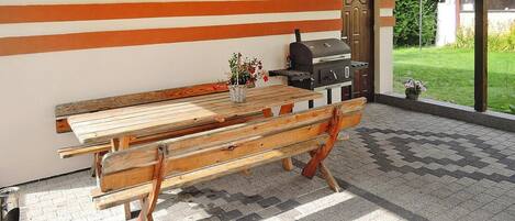 An outdoor patio with a wooden table and benches, a barbecue grill, and a view of a garden.