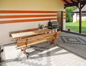 Um pátio externo com mesa e bancos de madeira, churrasqueira e vista para um jardim.
