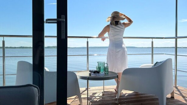 Una persona vestida de blanco de pie en una cubierta con vista a un cuerpo de agua tranquilo y una costa lejana.