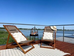 Two wooden deck chairs with cushions, a small table with a bottle and glasses, and a view of a lake.
