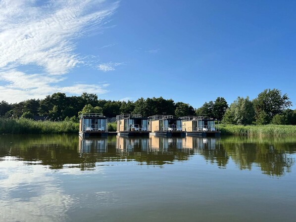Three floating structures with glass walls, situated on calm water, surrounded by greenery and a clear sky.