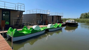 Una estructura flotante moderna con cubierta de madera y zona cubierta, amarrada junto a un río tranquilo.