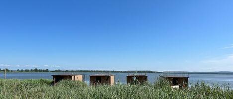 A waterfront property with three wooden cabins, surrounded by tall grass and a clear blue sky.