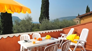 A terrace with a table set for two, two white chairs, a yellow umbrella, and a view of trees and hills. - Apartments Poiano in Garda (Garda)
