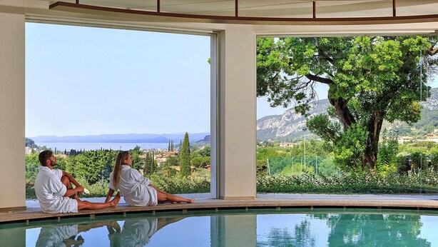 A pool with two people sitting by the poolside, a large tree, and a view of the landscape. - Apartments Poiano in Garda (Garda)