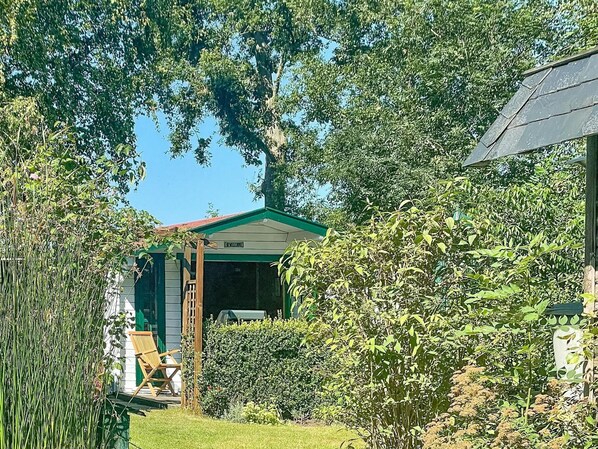 Una casa con techo verde, terraza de madera y jardín con diversas plantas y una silla.