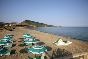 Une plage bordée de rangées de parasols verts et blancs, une rangée de chaises de plage et un petit bateau sur le sable.