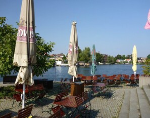 An outdoor seating area with tables and chairs by a waterfront, featuring umbrellas and a view of the water and buildings in the background. - Reihenhaus in Malchow (Malchow)
